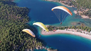 Drei Paragleiter schweben im Formationsflug über einer Bucht. Es sind Tandem-Gleitschirme. Das Wasser unter ihnen ist blau und klar. Der Strand lädt zum Sonnenbaden ein.