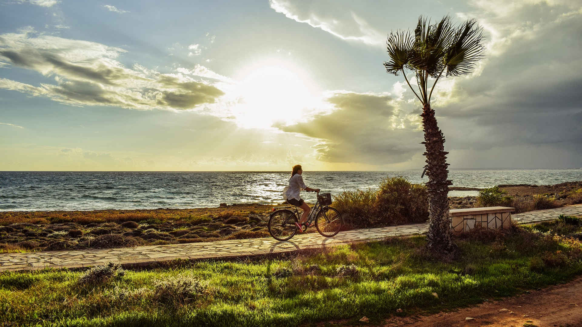 Eine Radwanderin fährt am Meer auf einem Weg entlang. Der Sonnenuntergang naht und es ziehen dunkle Regenwolken auf – Eine tolle Stimmung!