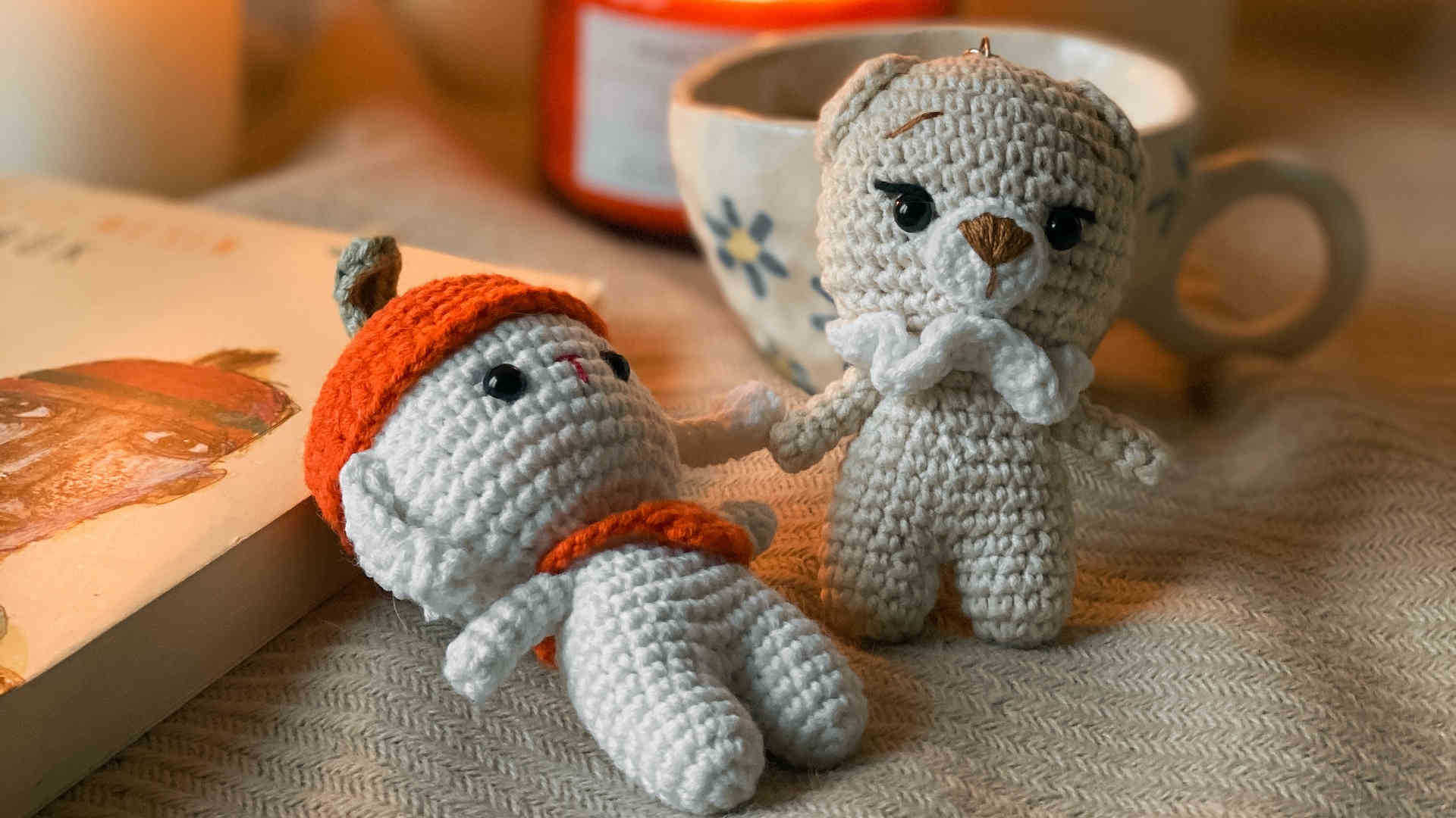 Wir sehen zwei niedliche Amigurumi-Figuren, die auf einem Tisch vor einer Kaffeetasse positioniert wurden. So wie es aussieht, können beide auch als Schlüsselanhänger verwendet werden.