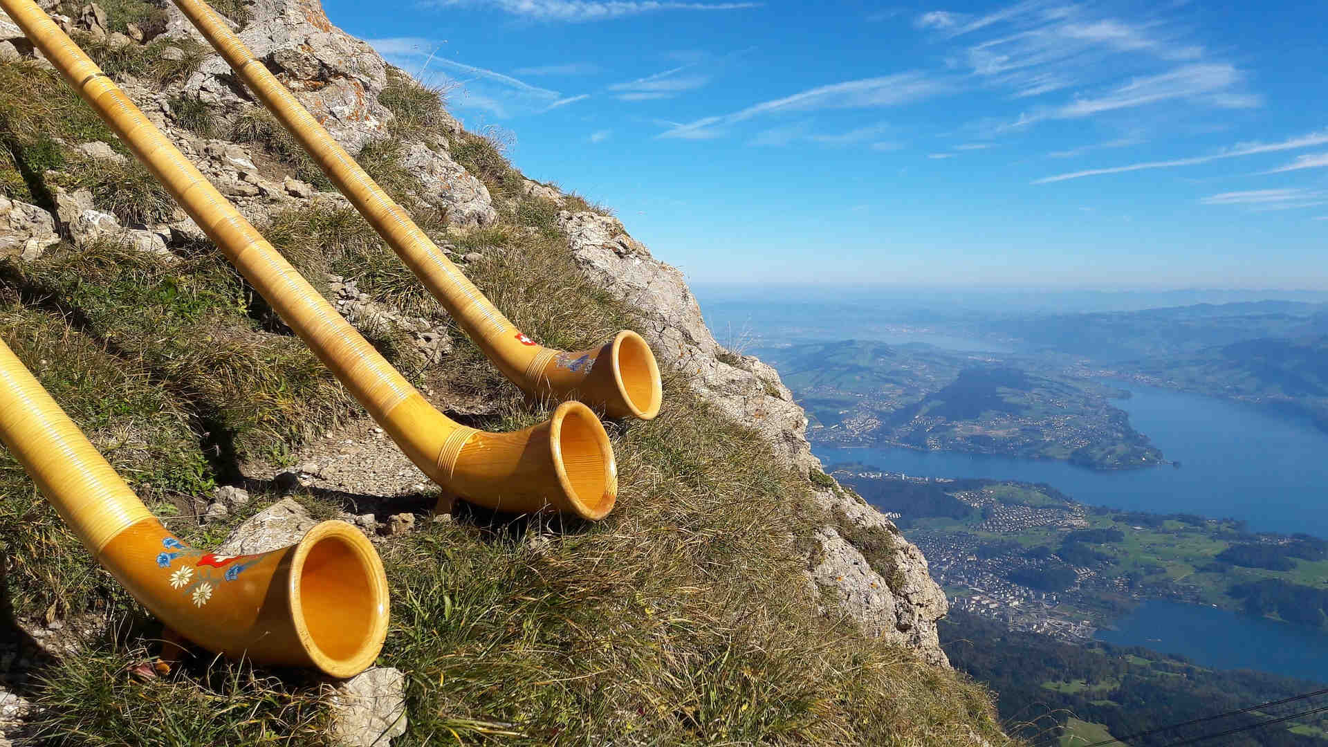 Drei Alphörner ruhen an einem steinernen Hang hoch über dem Tal mit Seen und Ortschaften. Vermutlich ist ihr Klang weit zu hören.